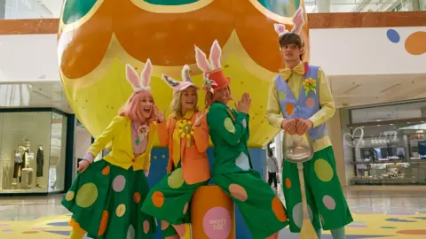 Four event workers are pictured wearing ears and bright costumes in front of a giant Easter egg
