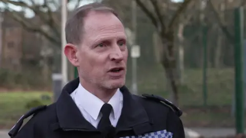 Ch Supt Simon Dodds gives a TV interview outdoors with trees and buildings in the background. He wears a police uniform and has receding grey-brown hair.