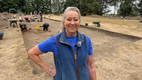Melanie, wearing a blue top and fleece vest, stands in the foreground of an active archaeological excavation site, where several people are carefully digging and examining large rectangular trenches in a dry, open field bordered by trees.