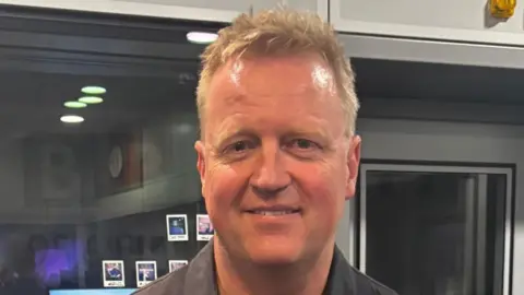 A photograph of a man wearing a black shirt looking directly into the camera. There is a glass window and door behind him.
