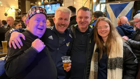 Three men and a woman stand smiling with their arms around each other in a crowded pub. A saltire hangs in the background and one of the men holds a beer