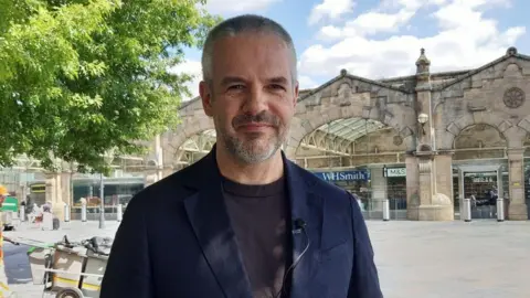LDRS South Yorkshire's Mayor Oliver Coppard - a man smiling at the camera with short grey hair n a navy suit jacket and jumper - standing in front of the stone-fronted Sheffield Train Station building with a tree on the left of the picture 