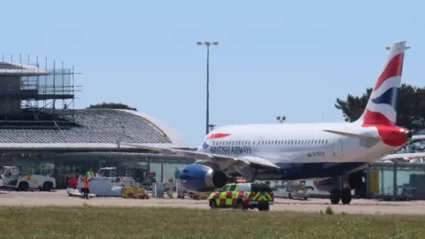 A view of a white red and blue British Airways aircraft in front of the airp[ort terminal building in Guernsey. 