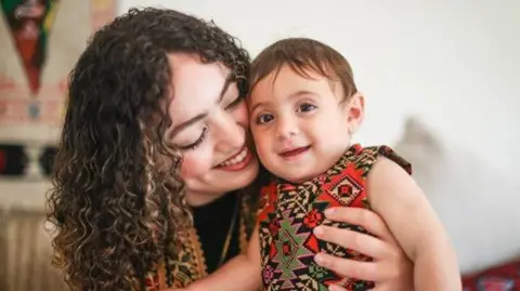 Hadil Louz, on the left, holding her daughter Laila. Hadil, who has long curly hair, is looking at her child, who is smiling at the camera and is wearing a patterned, sleeveless outfit.