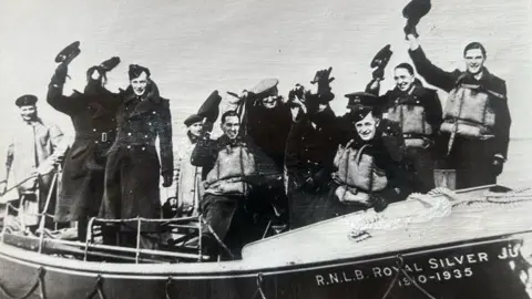 A black and white photo of the RNLI crews from WW2. They are in a lifeboat raising their hats in the air to wave. 