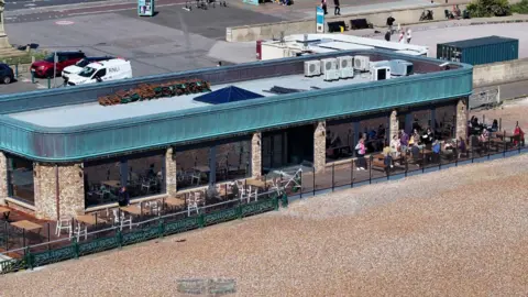 An aerial image of the cafe on Hove seafront. The roof is blue. There is seating on a terrace on the beach. 