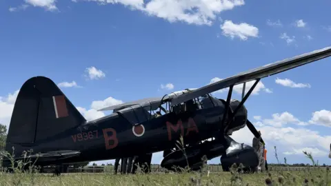 Jamie Niblock/BBC An old style aircraft that dates back to World War Two rests on a grass airfield on a sunny day. Some people can be seen admiring it from nearby.