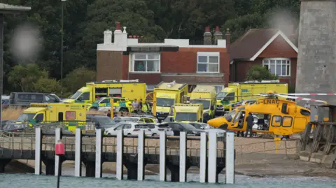 Ambulances, emergency response vehicles and an air ambulance helicopter are gathered near the seafront during an emergency operation.