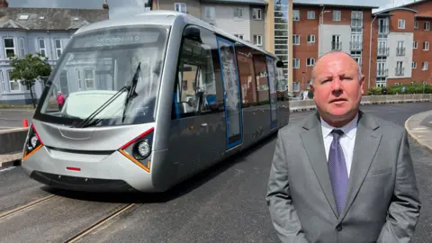 A man stands in front of the Very Light Rail (VLR) tram vehicle on tracks in Coventry city centre. He is wearing a grey suit and purple tie. A modern-looking grey carriage is riding past on the demonstrator track behind him as part of testing.
