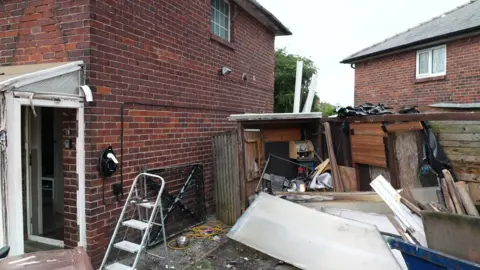 The side of a semi-detached house with fire damage to the white porch and shed. There is waste strewn across the garden.