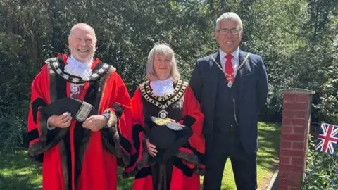Two men and a woman are standing in a garden. The man and woman on the left are wearing ceremonial robes and  the man on the right is wearing a suit and ceremonial chain.