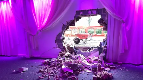 An image taken inside Arleston Hall. Purple lights colour the white walls. A large hole is visible in the middle of the wall, with a wheelbarrow visible outside. 