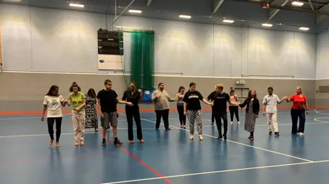 Group of people holding hands and taking part in an activity on an indoor sports hall court.