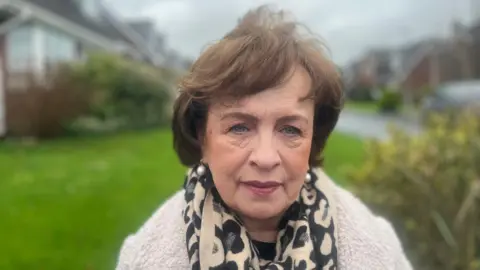 Dianna Dodds wearing a cream knitted top, pictured in front of a green lawn and buses. She has dark hair and a leopard print scarf