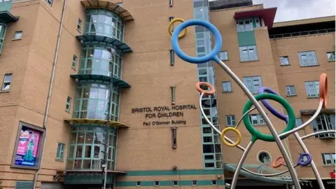The front of the Bristol Royal Hospital for Children building from the street. The building is several storeys high, and the exterior is of light brown brick. There is a piece of colourful artwork in front of it, made of different coloured hoops attached to the ends of curved metal poles