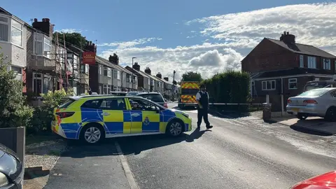 BBC Police officer stands in the road in front of police tape which is cordoning off an area surrounding a house on a residential street. A police car is parked in front of the cordon with a police van inside the police cordon.