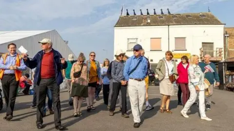 A crowd of people taking part in a guided walk. There is a white building in the background with boarded-up windows.