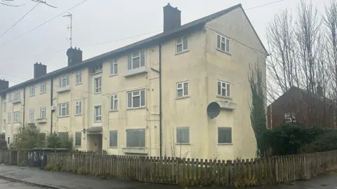 A block of flats painted white. Most of the windows on the ground floor of the three-storey building are boarded up. There is a low fence made of vertical wooden slats next to the pavement around one end of the block. 