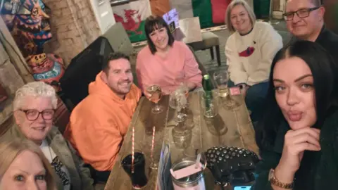 Supplied Seven people sitting around a table in a pub. There are several drinks on the table and the flags of several nations attached to the wall behind them.