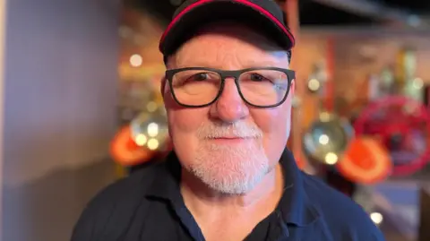 Dave Farries is looking at the camera. He is wearing black-rimmed glassed and has a grey goatie beard. He had a fire service cap on his head with a crest on the front. The Museum of Scottish Fire Heritage is behind him but it is blurred in the image.
