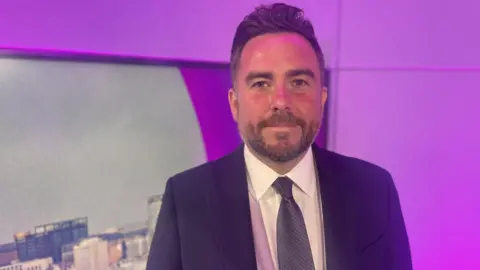Portrait of a person wearing a dark business suit, white shirt and patterned tie, standing indoors in front of a purple-lit backdrop. A cityscape image is partially visible on the left, while the background features modern wall panels illuminated with pink and purple lighting. The person is centred in the frame and facing the camera.