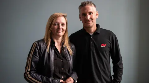 PA Adam Staples and Lisa Grace smile as they stand against a grey background. Lisa has long, straight blonde hair parted to the side and is wearing a black leather jacket with gold stripes down the sleeves, while Adam, who has short, grey hair, is wearing a long-sleeved black polo shirt with a red and white logo on the breast.