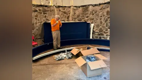 Friends of Newchapel Observatory James is stood inside the building looking down at items in a pile on the floor with his hand on his chin. He has an orange t-shirt on and brown trousers. He is stood in a circle and boxes on the floor also contain various items.