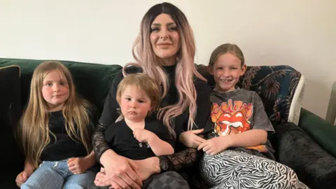 A woman is sitting on a sofa with a small boy on her lap and two young girls sitting either side of her. 