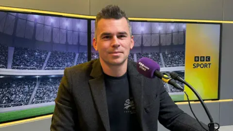 A man in a suit jacket over a dark coloured top. He is sitting in front of a microphone, with BBC Sport branding behind him, with an image of a crowd in a stadium.