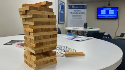 Emma Baugh/BBC A stack of wooden jenga sticks piled in a neat tower, on a white round table in a conference room. There is another white table in the distance in front of a sign saying youth trailblazer. A large TV screen is hanging on the wall.
