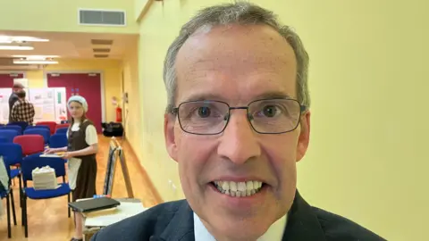 Mr Paisley wears black rectangular glasses and has short grey hair. He smiles at the camera as he wears a black suit jacket and a white shirt. He has brown eyes and is standing in front of a yellow wall, in the background is a young girl wearing a brown dress and white hat standing next to a blue chair.