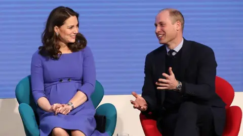 Catherine wearing a blue Seraphine dress, sitting on a turquoise chair, looking at Prince William sitting on a red chair next to her, who is talking and gesturing with his hands.