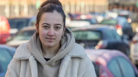 Stockport resident Hannah Miles wearing a beige hoodie and jacket stood outside Meadow Mill in a car par.