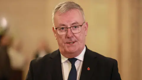 PA Health Minister Mike Nesbitt wearing a dark suit with a poppy badge on the lapel. There is a blurred background. 