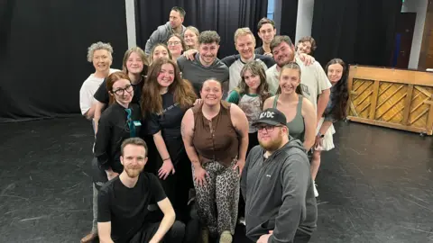 A large group of people gathered in a theatre rehearsal space with black curtains and stage lighting. Some people are standing while others are kneeling in front.