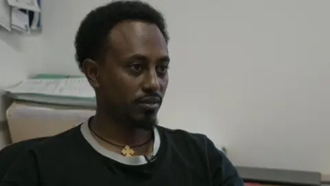 Sam with black hair and beard wearing a black and grey top and a wooden cross necklace sits in an office with paperwork behind him. He looks impassively away from the camera.