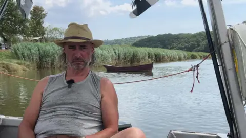 Will is wearing a hat and grey T-shirt as he sits on the ferry with the river behind him.