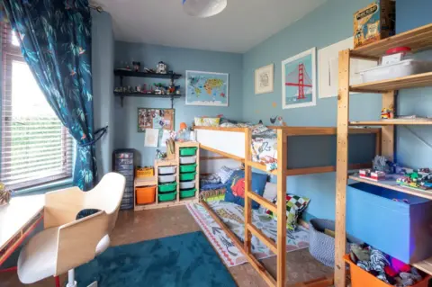 IWC Media Inside Homegrown Hoose. A children's bedroom with wooden furniture, toys on shelves and posters on the wall. The walls are painted blue and there are colourful rugs on the floor.