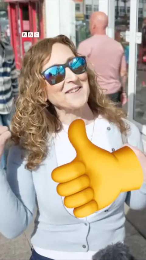A woman wearing sunglasses smiles at the camera, there is a big thumbs up emoji in front of her