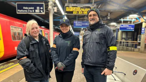 Lisa Townsend stands in Guildford railway station, flanked by two members of the Street Angels. All three are wearing branded jackets.