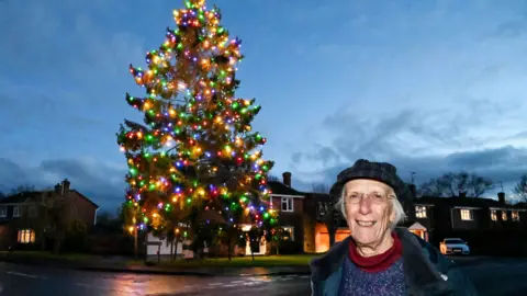 SWNS An elderly woman with white hair wears a dark coloured hat, green coat, purple jumper and red neck warmer while standing in front of a large Christmas tree that has been illuminated with multi-coloured lights. 