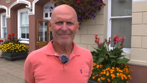 A man is pictured stood outside a parish hall. There are colourful plants visible behind him, with orange and yellow flowers. He is bald and wearing a coral coloured polo shirt. He his smiling somewhat. 