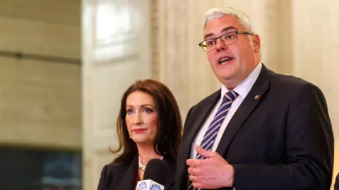 PA Media DUP Deputy First Minister Emma Little-Pengelly with party leader Gavin Robinson in the Great Hall at Stormont Great Hall.
On the right - Gavin Robinson - a tall man with grey hair and glasses. He is speaking into a microphone and is wearing a suit and tie,
On the left - Emma Little Pengelly has long dark brown hair, she is wearing a dark coloured jacket and a pearl necklace