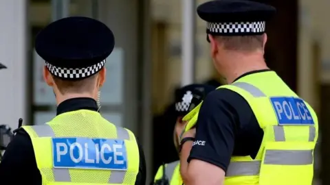 The backs of two police officers, both with short dark hair under black caps with a black and white checkered headband. They wear green high-vis waistcoats with "police" written on the back.
