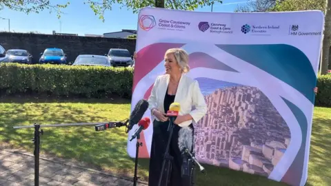 First Minister Michelle O'Neill, standing in front of three microphones, outdoors. She has blonde hair, tied up, and is wearing a black dress and a white blazer. There is a large poster behind her with an image of the Giant's Causeway on it. 