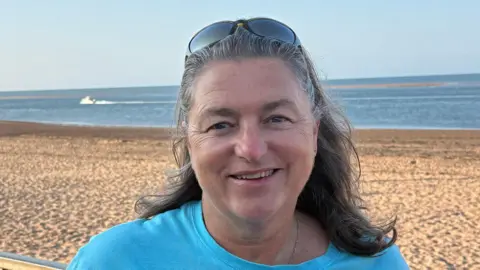 Jill Gathercole she has shoulder length dark hair and sunglasses on he rhead. She is stood in front of a beach. She is wearing a blue t shirt. 