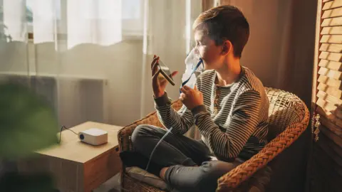 A boy in a striped T shirt sitting on a rattan chair holds a mobile phone while breathing through an inhaler