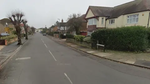 A residential tree-lined street with houses on either side and a car is parked on the right-hand side. 