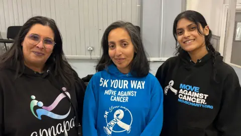 Image shows Indy Samra on the left from Apnapan wearing a black hoodie that says Apnapan on it. She's wearing tinted glasses and has long black hair. Her arm is around Nazeem Bi, in the middle who is wearing a blue hoodie which says 5k your way move against cancer on it, with the move against cancer logo on it. She also has black hair but that is shoulder length. On the right is dance instructor Janyna Patel who is wearing a black hoodie which says move against cancer.movementcharity.org on it. She has long black hair which is tied into pigtails. 