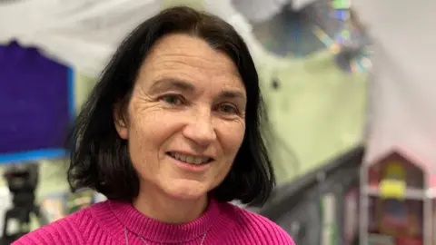 Joan Henderson with shoulder length black hair is smiling at the camera as she wears a pink top.